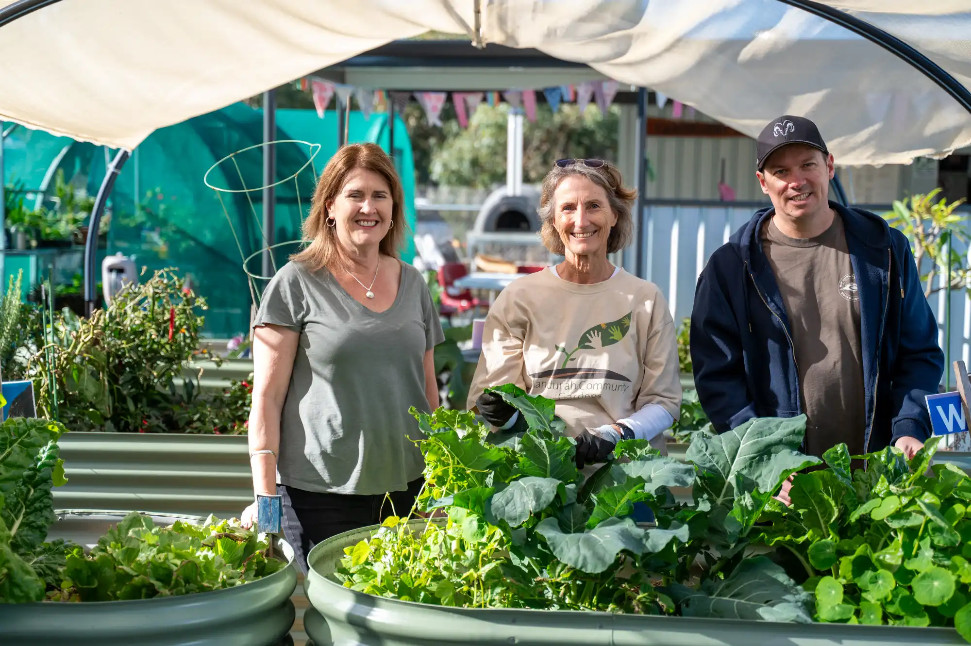 Three people stand among raised vegetable garden beds beneath a shade structure, holding freshly picked greens. Lush leafy crops, garden equipment and community garden facilities surround them.